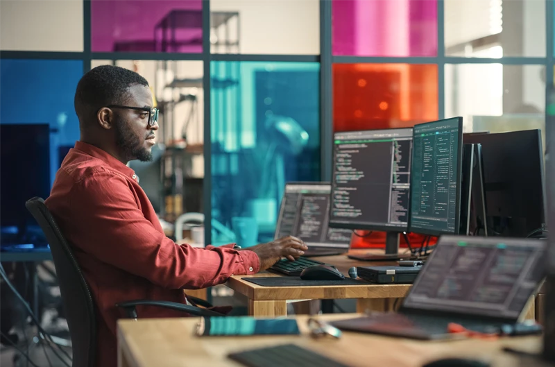 African American Man Writing Lines Of Code On Desktop Computer With Multiple Monitors and Laptop in Creative Office. Male Data Scientist Working on Innovative Online Service For Start-up Company.